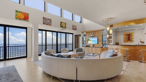 A spacious living area with a curved white sofa, a glass coffee table, and a large TV. The room has a balcony with a view of the ocean. - Penthouse on the Beach! Beautiful Sunset Views! (Fort Myers)