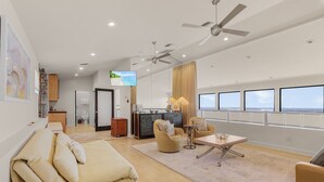 A spacious living area with a sofa, armchairs, a coffee table, and a TV. There are ceiling fans, a kitchen area, and a view of the ocean through large windows. - Penthouse on the Beach! Beautiful Sunset Views! (Fort Myers)
