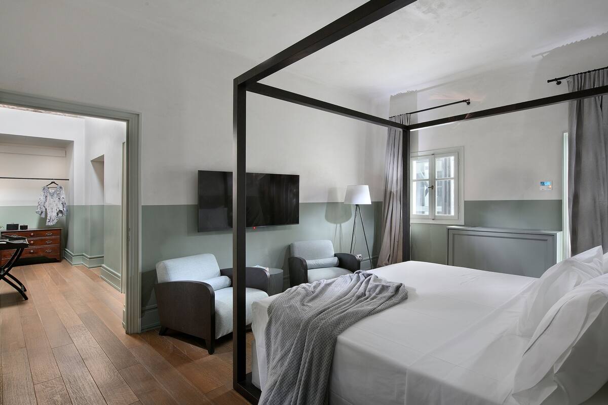 Une chambre d’hôtel moderne dotée d’un lit à baldaquin, d’un bureau avec une chaise, d’un téléviseur à écran plat et d’une grande fenêtre avec des rideaux.