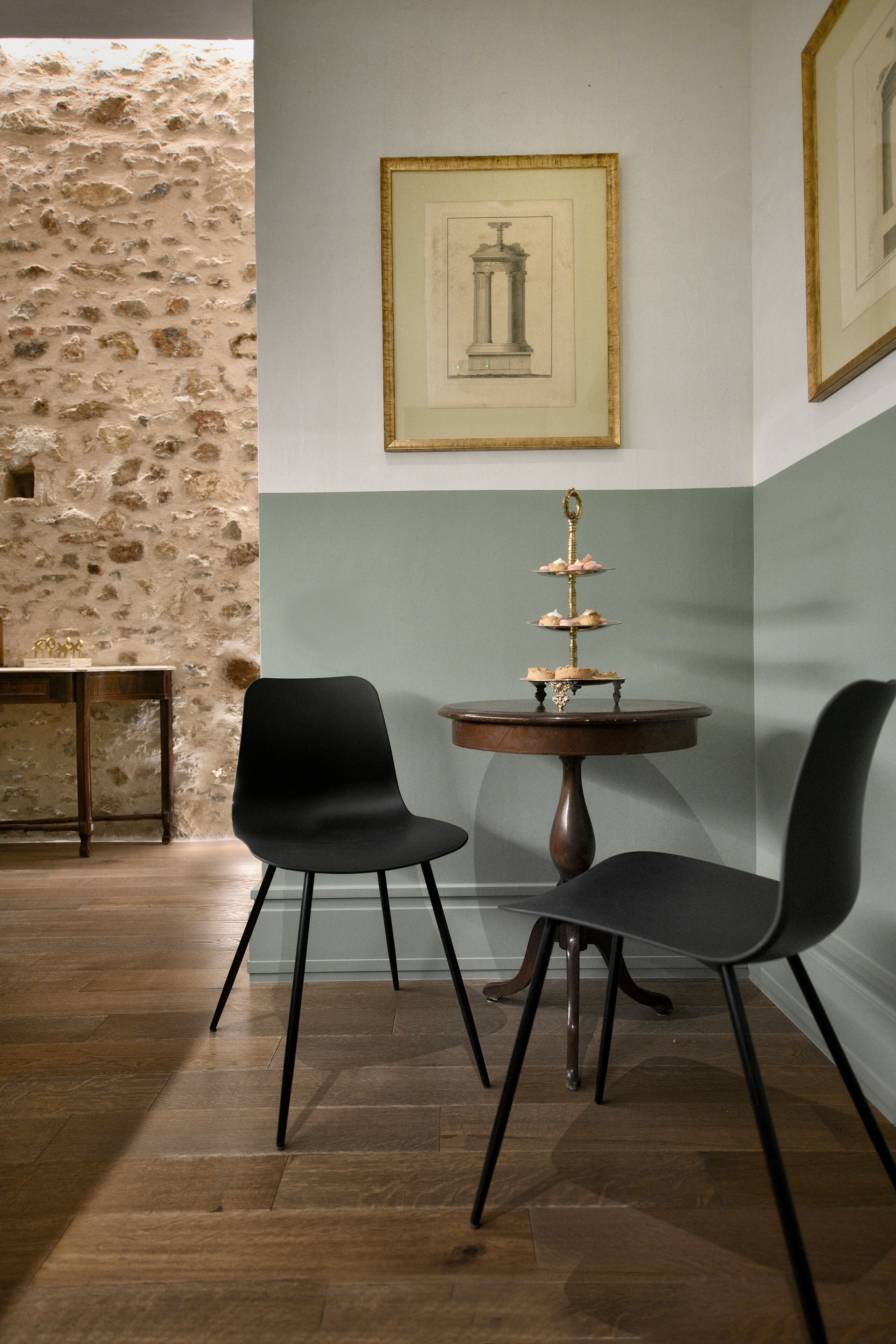 Une pièce avec un mur en pierre, deux chaises noires, une table ronde surmontée d’un présentoir à desserts et des œuvres d’art encadrées accrochées au mur.