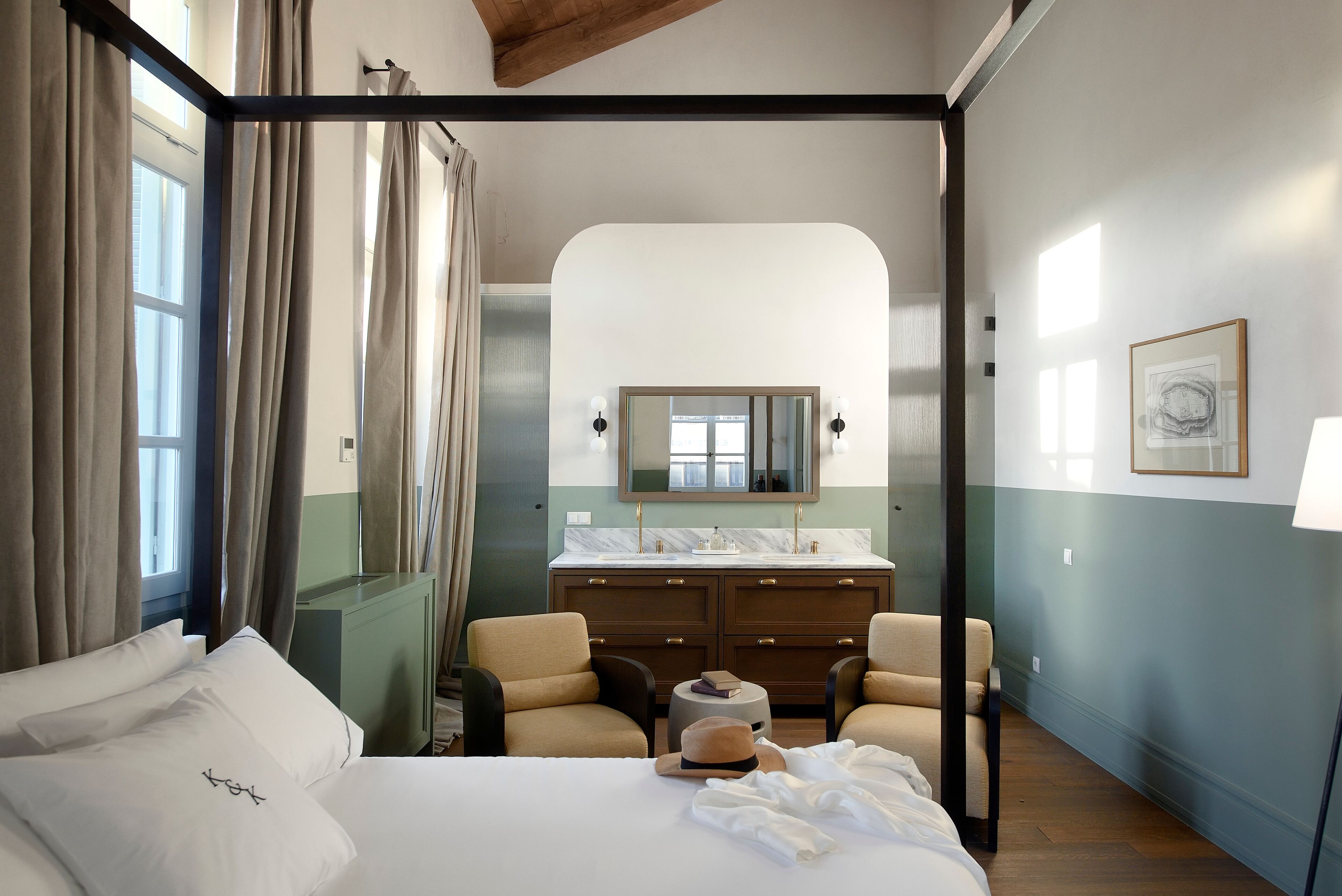 A hotel room with a bed, two armchairs, a vanity with a mirror, and a window with curtains.