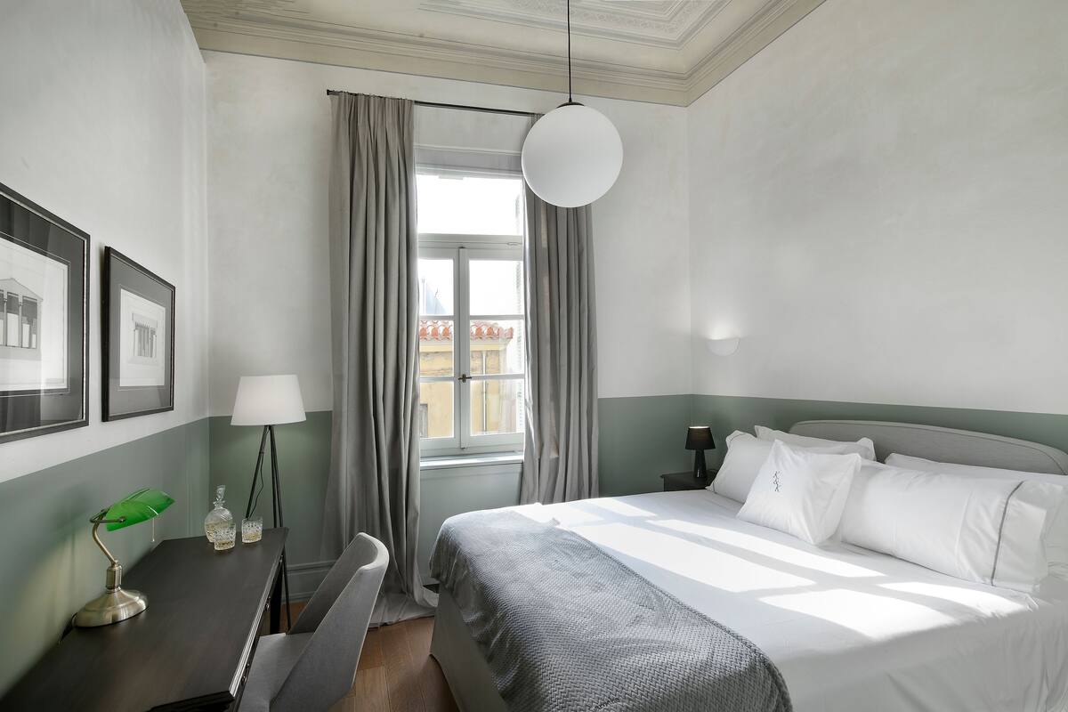 Une chambre d’hôtel avec un lit, un bureau doté d’une lampe, une chaise, une fenêtre avec des rideaux et des tableaux encadrés accrochés au mur.