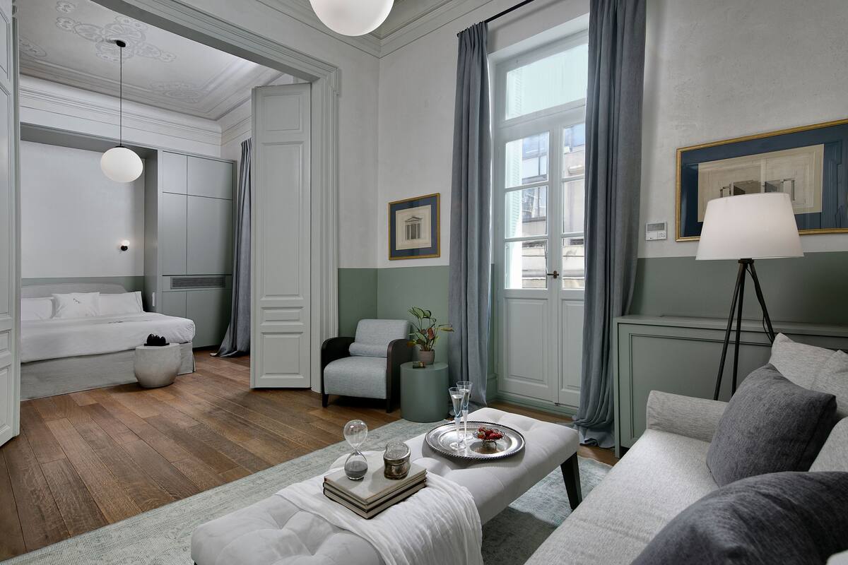 Un salon moderne comprenant un canapé, une table basse, une table d’appoint et un fauteuil. On y trouve une fenêtre avec des rideaux, une lampe et un tableau encadré au mur.