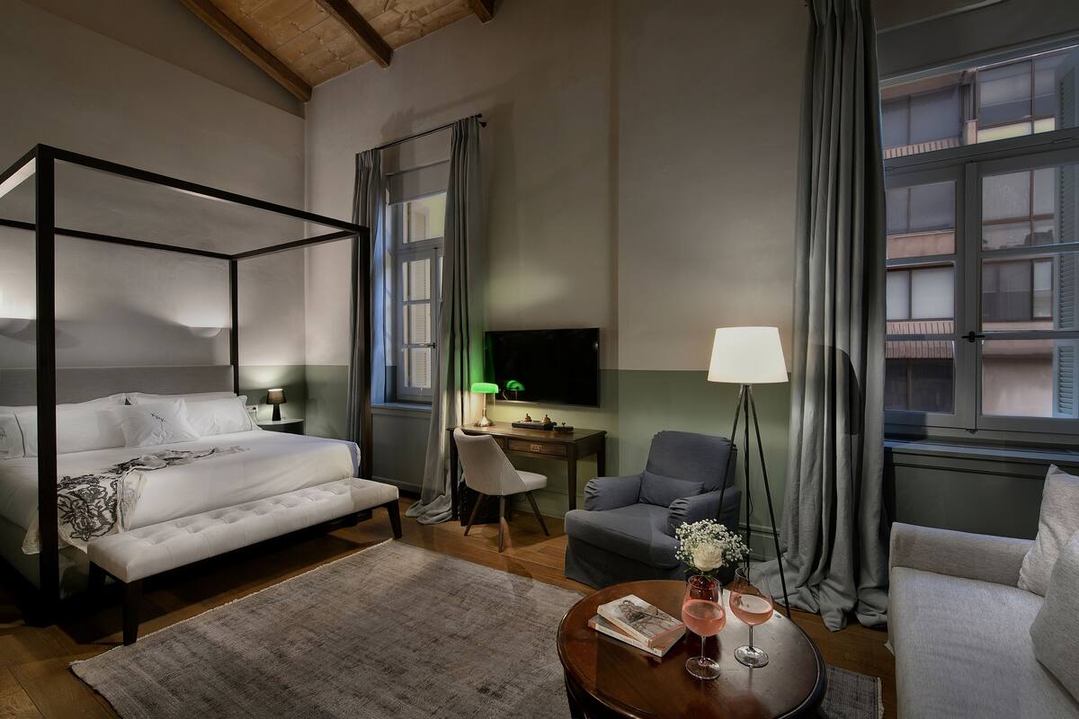 Une chambre d’hôtel bien éclairée, avec un lit à baldaquin, un bureau avec un ordinateur, un fauteuil gris et un canapé.