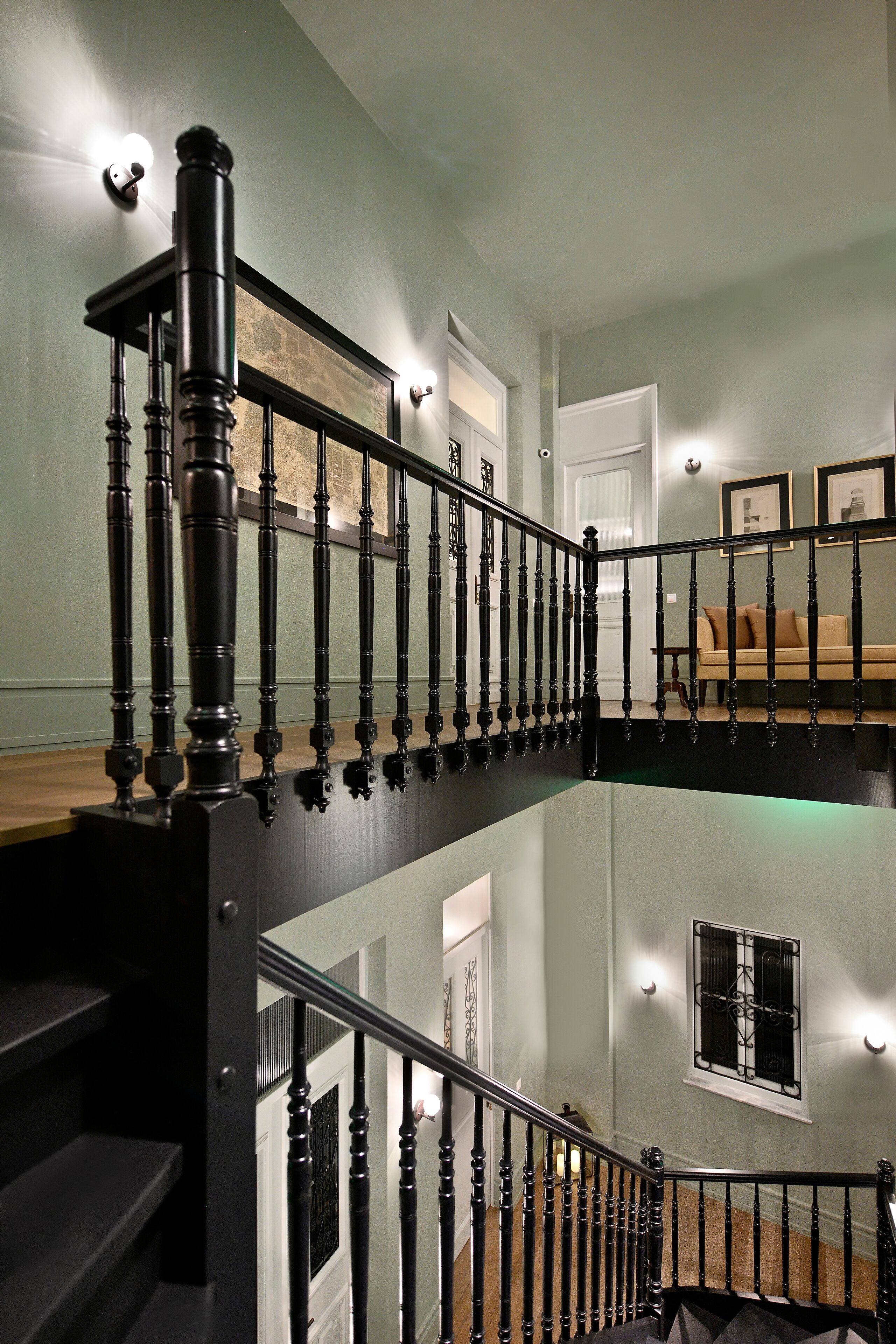 Un escalier à plusieurs niveaux, doté de rampes noires et d’une finition en bois, mène à un palier où se trouvent un canapé beige et une fenêtre ornée de barreaux en fer décoratifs.