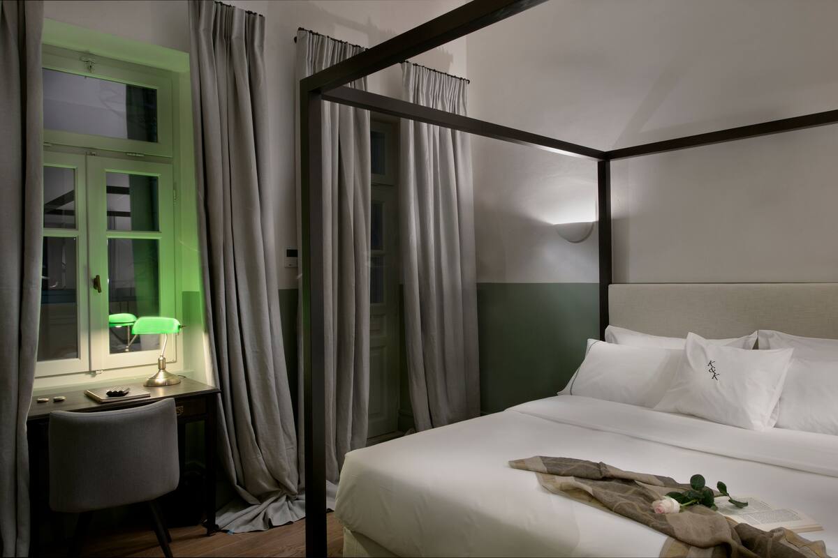 Un lit à baldaquin avec du linge de lit blanc, un bureau avec une lampe et une fenêtre avec des rideaux.