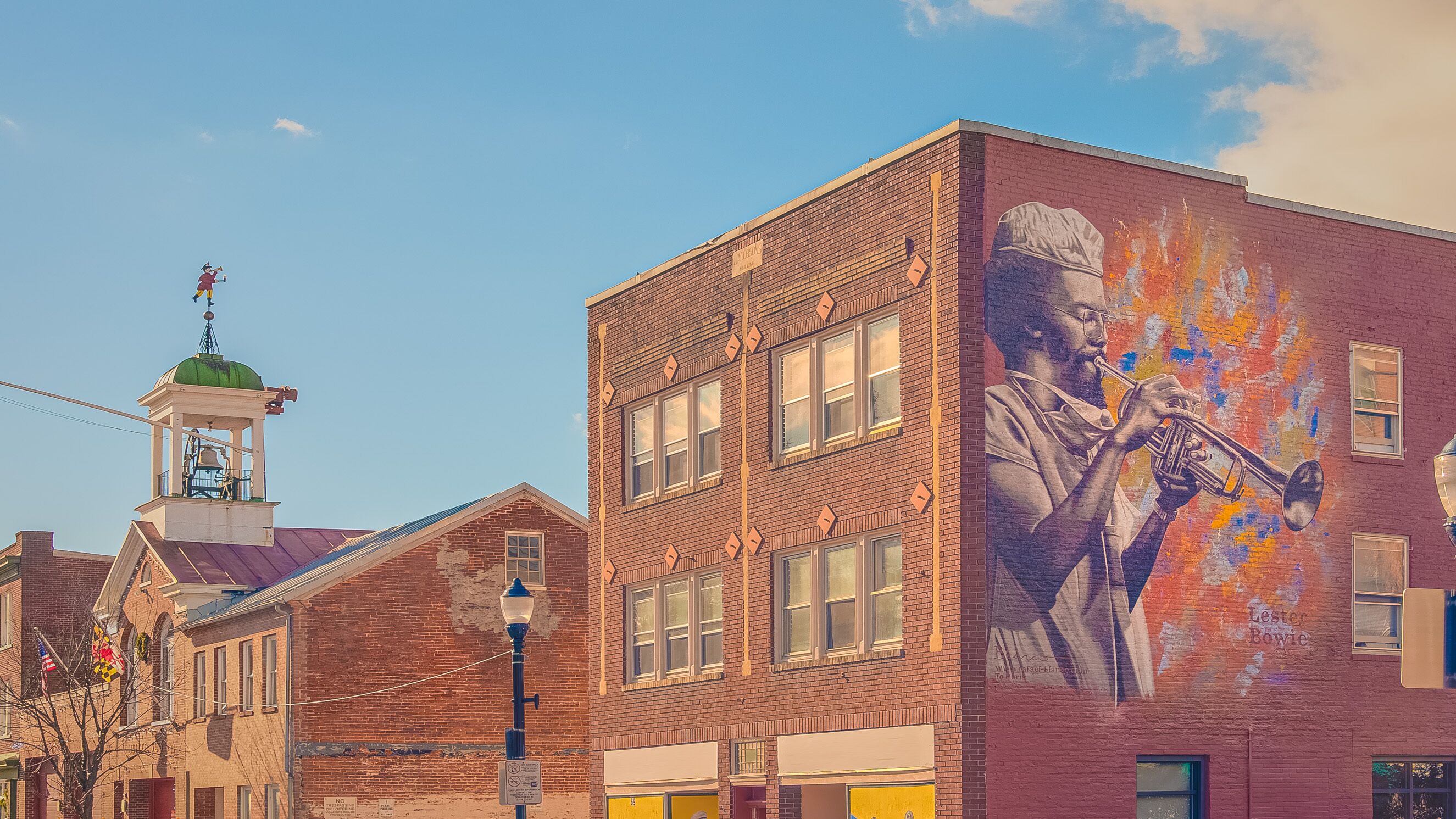 A building with a mural of a man playing a trumpet, a clock tower, and a street with a no parking sign.