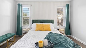 A neatly arranged bedroom with a bed, green curtains, a bench, and a tray with two cups. - Emerald Indigo House | Pet Friendly (Dallas)