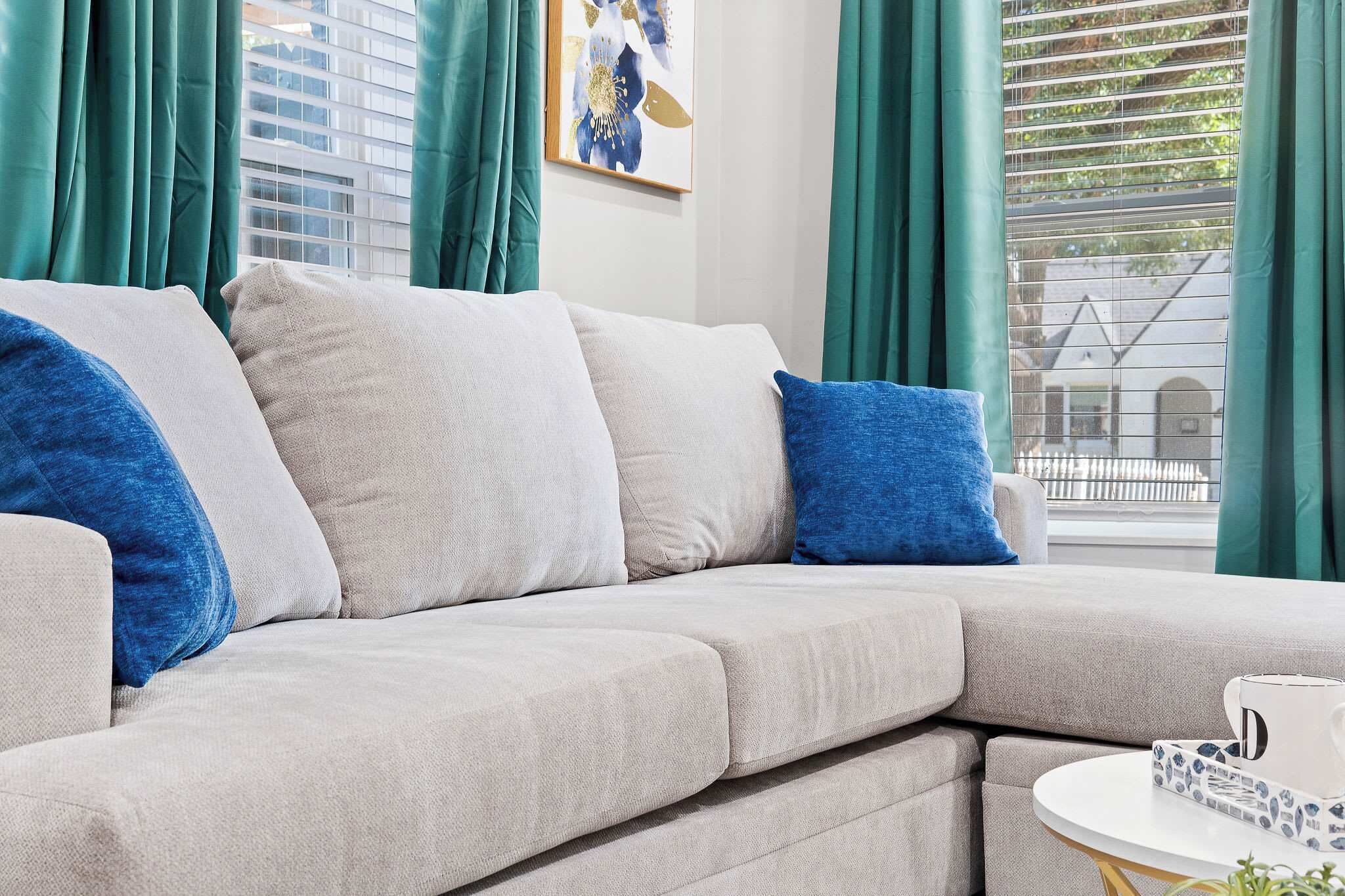 Una sala de estar moderna con un sofá gris, cojines azules, una mesita redonda con una taza blanca y una ventana con cortinas verdes.