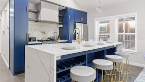 A modern kitchen with blue cabinetry, a white marble island, and a large window with blinds. - Emerald Indigo House | Pet Friendly (Dallas)