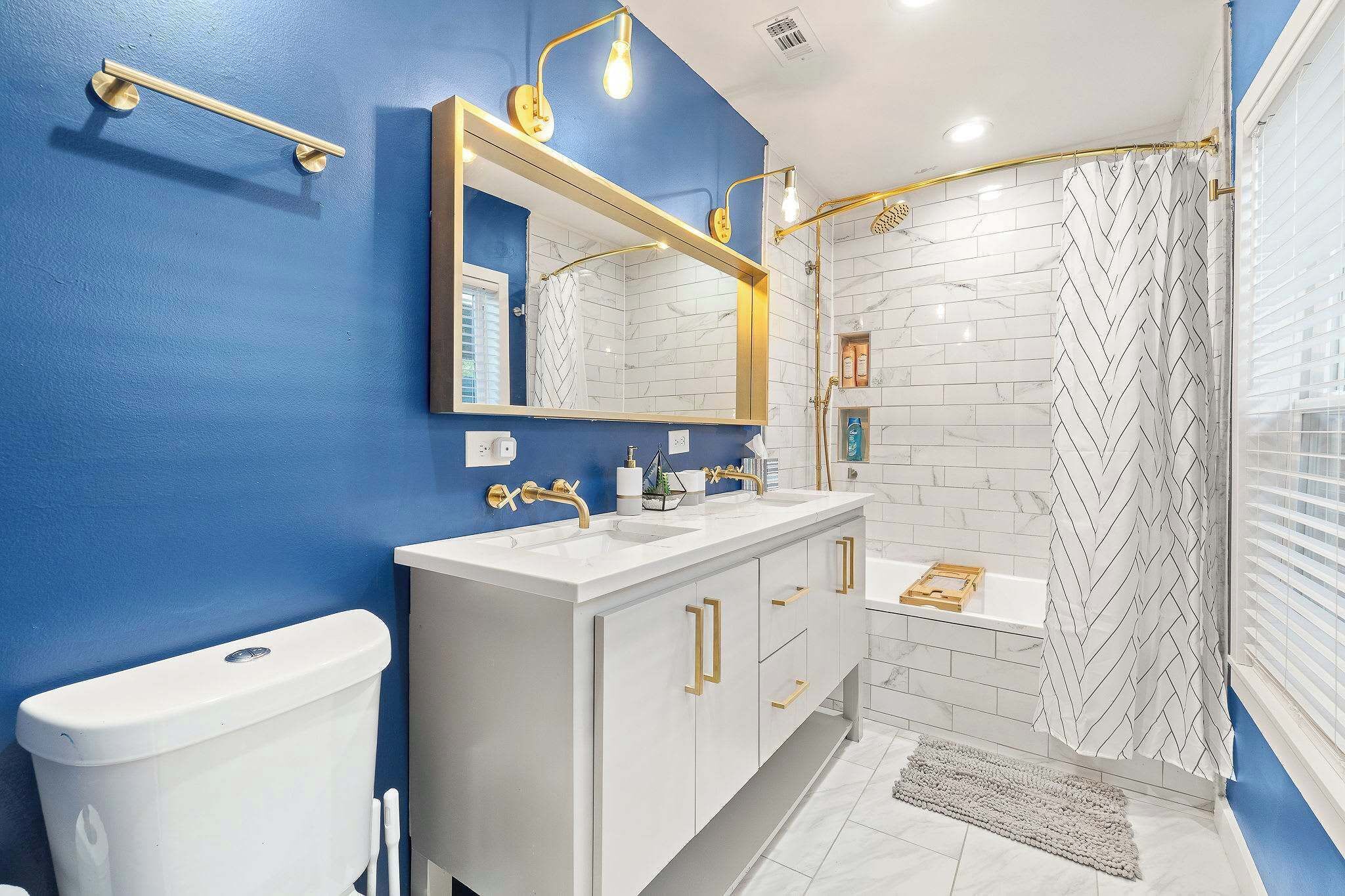 A bathroom with a blue wall, white tiled shower, and a white sink vanity with gold handles.
