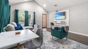 A modern living room with a dining area, a TV mounted on the wall, and a view of the outdoors through the windows. - Emerald Indigo House | Pet Friendly (Dallas)