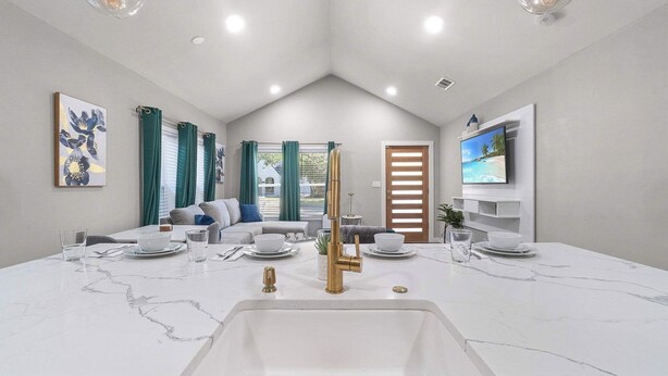 A modern kitchen with a white marble countertop, gold faucet, and a built-in sink. There is a dining area with a table set for two, and a living room with a sofa and a TV. - Emerald Indigo House | Pet Friendly (Dallas)