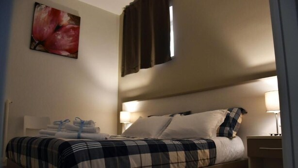 A neatly made bed with a checkered bedspread, two bedside lamps, and a wall-mounted painting of flowers. - Villa 18 - One-Bedroom House (Matera)