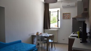 A compact room with a blue sofa, a dining table with chairs, a kitchen area with a stove and sink, and a window with curtains. - Villa 18 - One-Bedroom House (Matera)