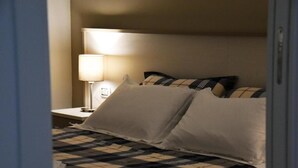 A neatly made bed with a checkered bedspread and white pillows, a bedside lamp, and a nightstand with a lamp. - Villa 18 - One-Bedroom House (Matera)