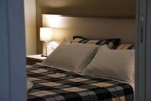 A neatly made bed with a checkered bedspread and white pillows, a bedside lamp, and a nightstand with a lamp. - Villa 18 - One-Bedroom House (Matera)