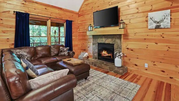 A cozy cabin interior with a stone fireplace, leather sofa, and a television mounted above it. - Family Cabin Fun: Deck Slide, Hot Tub, Fire Pit & Resort Amenities! (Ellijay)