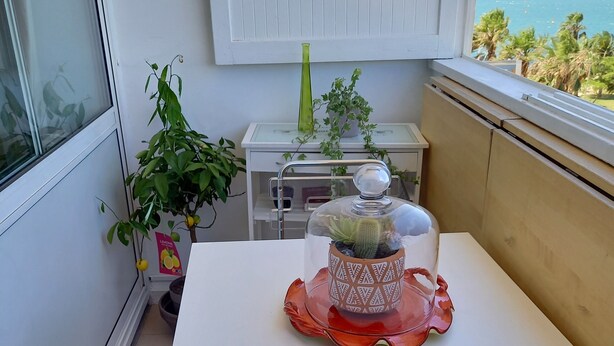 A balcony with a white table, a clear glass chair, a potted plant, a glass container with a lid, and a view of the sea. - La Goelette 4 (Toulon)