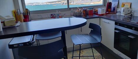 A kitchen with a view of the sea, a dining table, chairs, and a refrigerator.