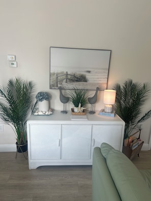 A living room with a white cabinet, a beach-themed painting, and two potted plants.