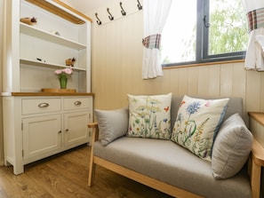 A cozy room with a wooden floor, a white cabinet with shelves, a grey sofa with floral cushions, and a window with curtains.