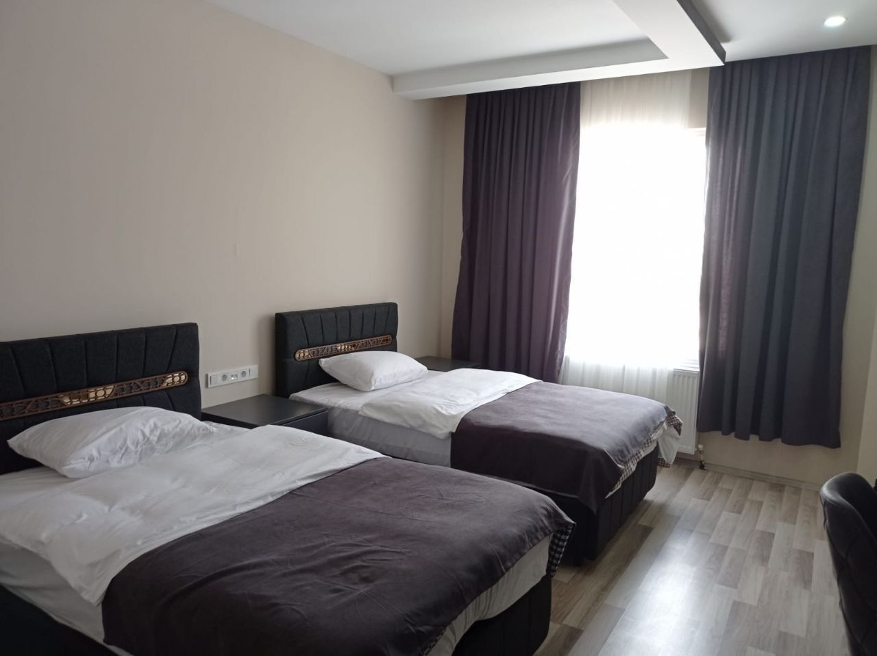A hotel room with two beds, a wooden headboard, a window with curtains, and a radiator.