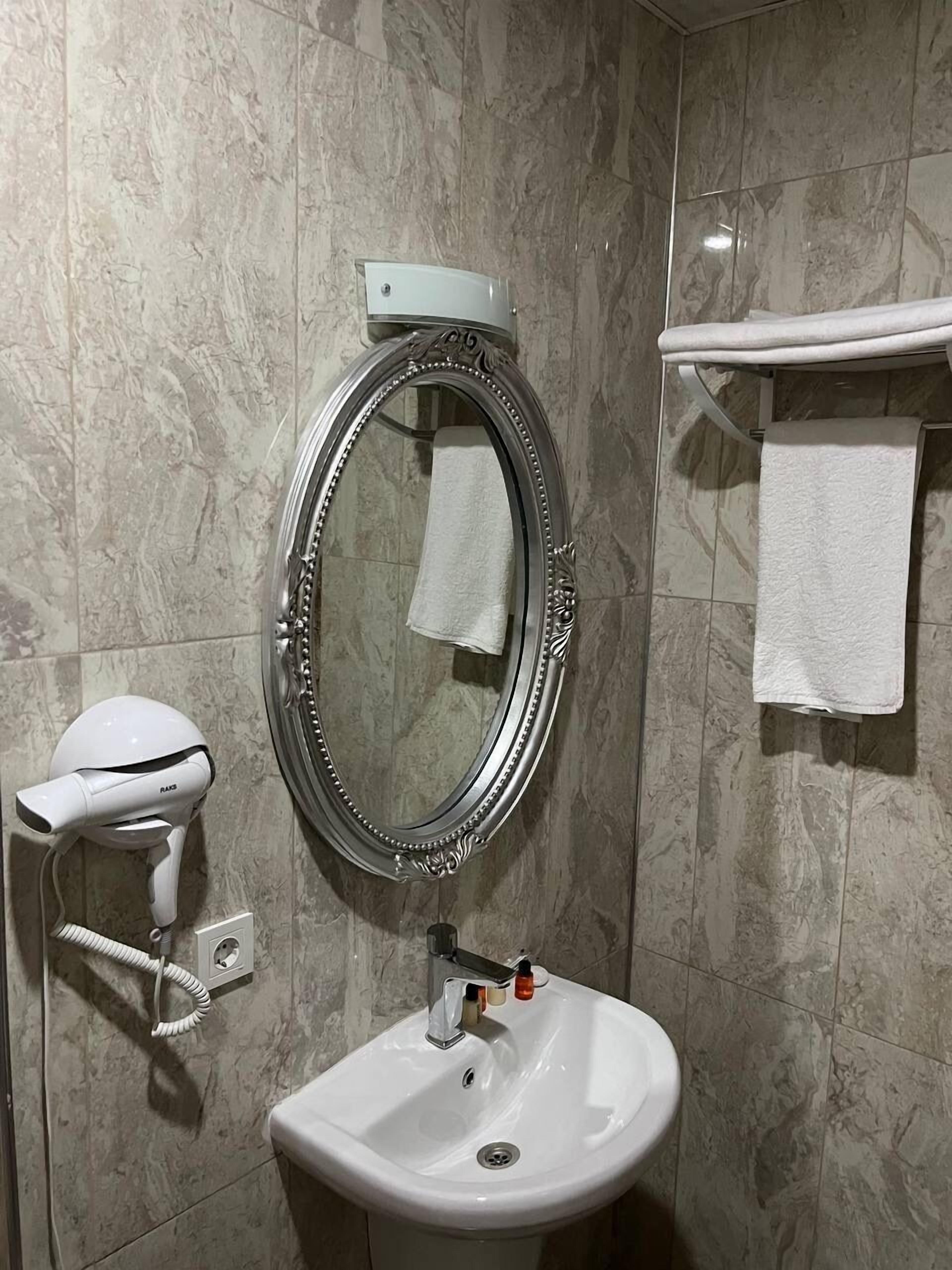 A bathroom with a marble wall, a round mirror with a decorative frame, a white pedestal sink, a handheld hair dryer, and a towel rack with two towels.