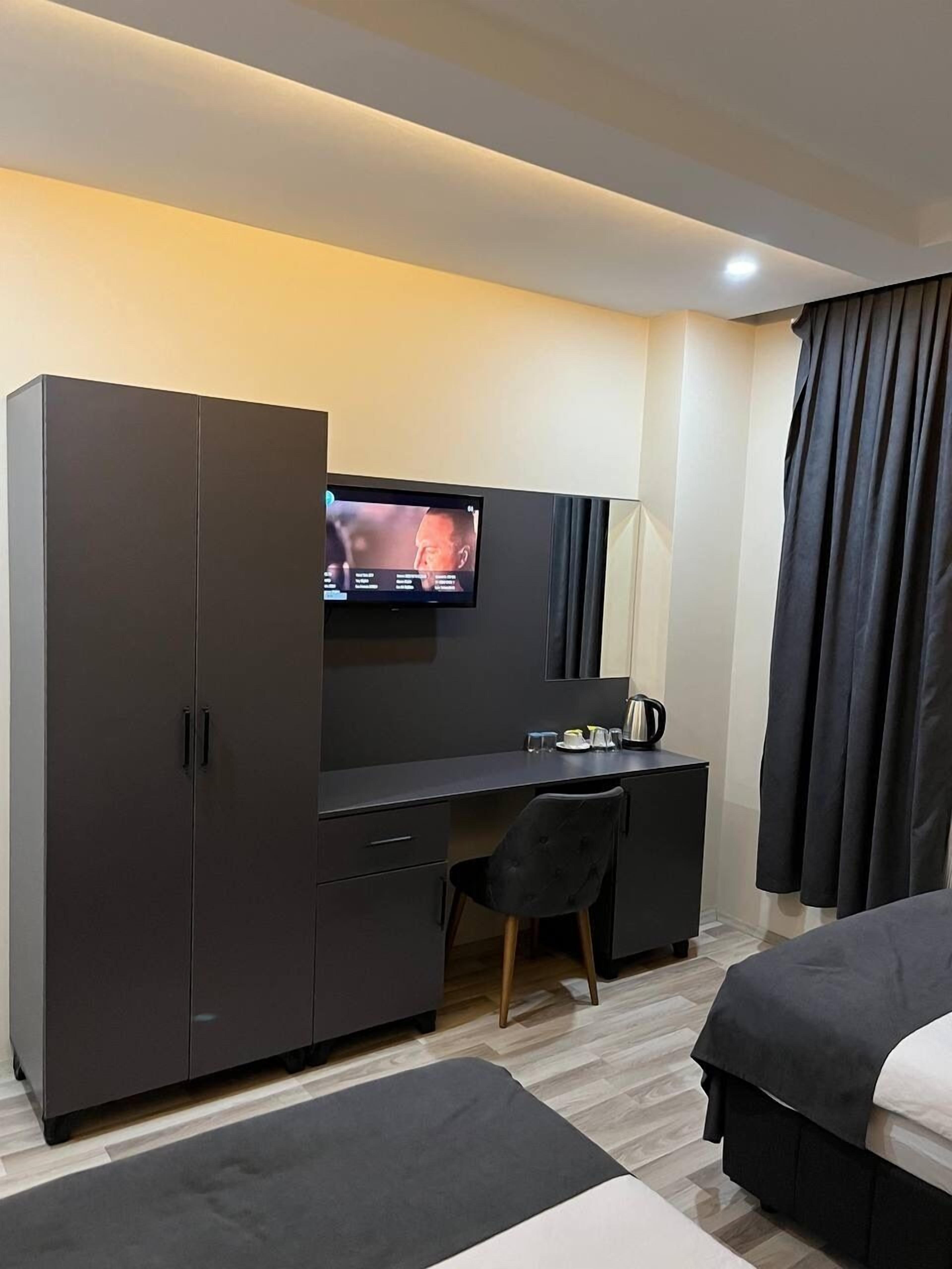 Une chambre d’hôtel moderne avec une armoire gris foncé, un bureau doté d’un téléviseur à écran plat, une chaise noire, un lit et une grande fenêtre avec des rideaux.