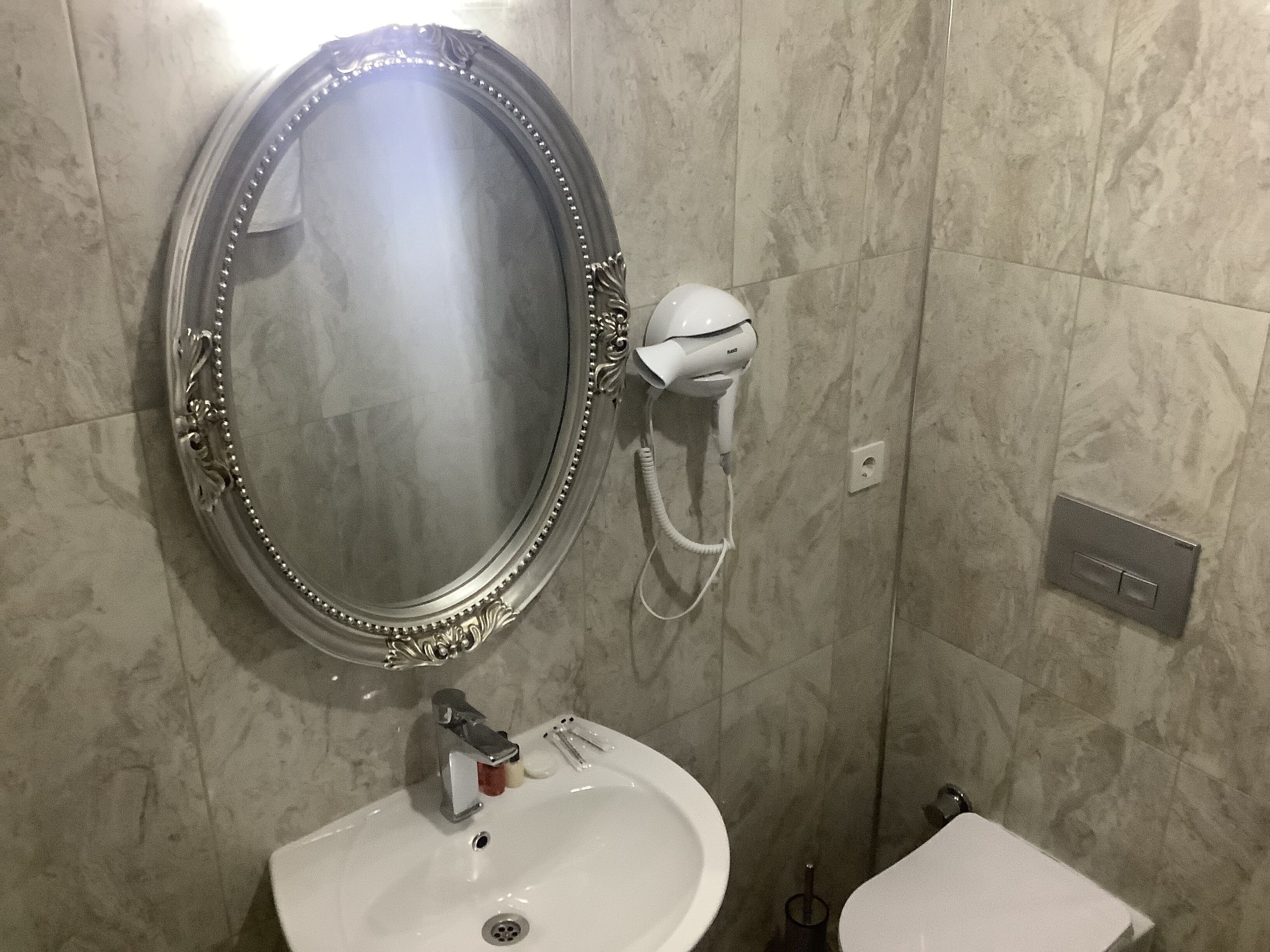 A bathroom with a marble wall, a round mirror with a decorative frame, a white sink, and a toilet.