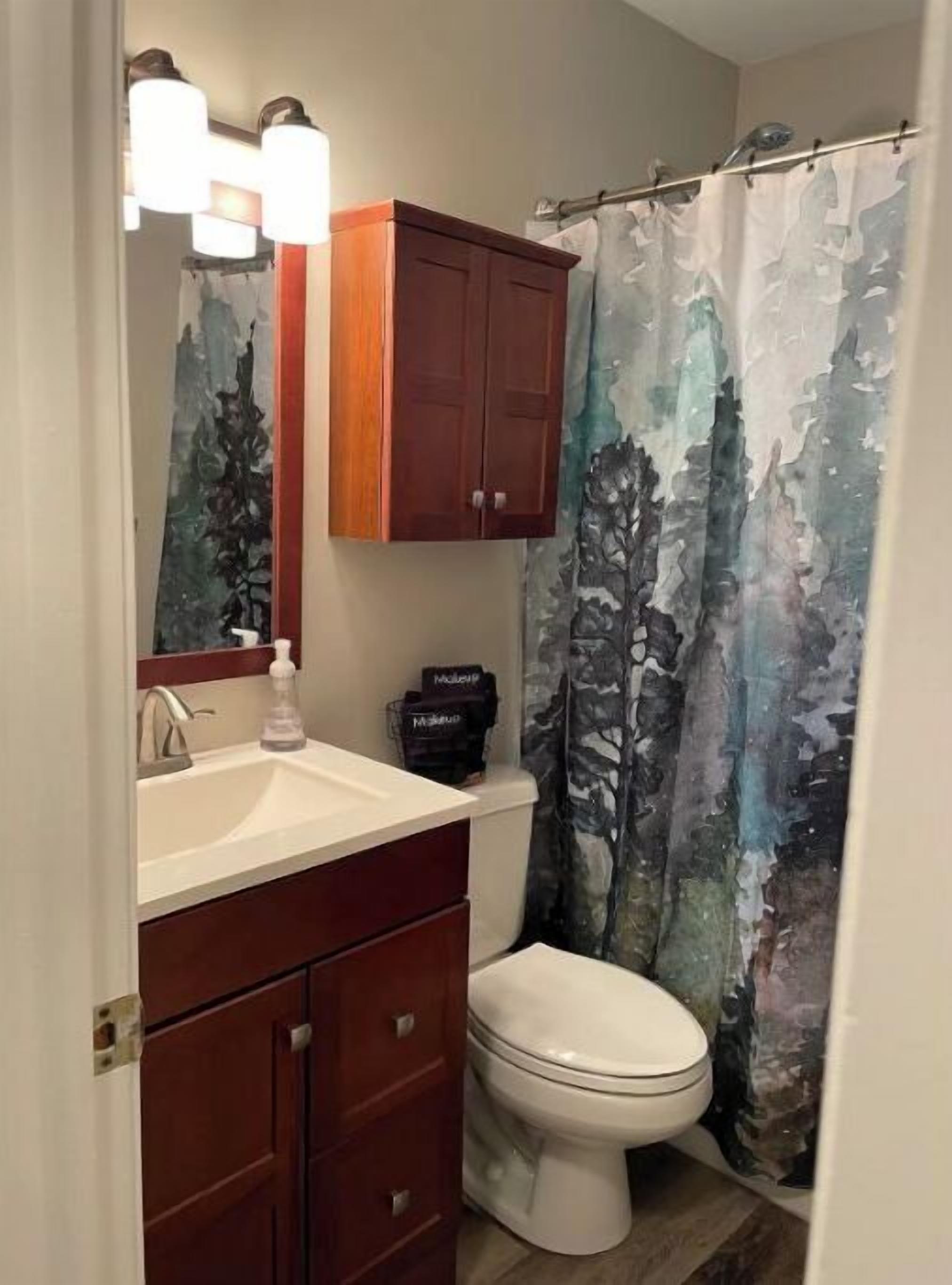 A bathroom with a white sink, wooden cabinets, a toilet, and a shower curtain with a forest pattern.