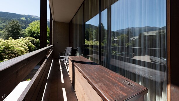 Ein Balkon mit einer hölzernen Bank und Stühlen, der auf eine gebirgige Landschaft mit großen Fenstern blickt.