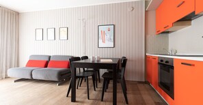 A modern living room with a grey sofa, red cushions, a black dining table, and chairs. There are framed pictures on the wall, a kitchen with orange cabinets, and a window with curtains.