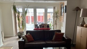 A living room with a dark leather sofa, a glass coffee table, and a red plaid tablecloth. There are two potted plants, a wooden cabinet, and a floor lamp. - Quiet rooms in a terraced house easy walking to shops, train (Birmingham)