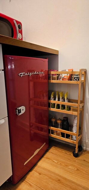 Kompakti keittiö, jossa on punainen Frigidaire-jääkaappi, puiset hyllyt, joilla on ruokatarvikkeita, ja mikroaaltouuni ylähyllyllä.