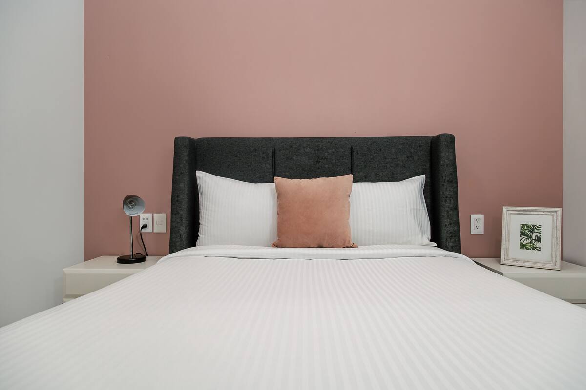 Una cama bien hecha con una colcha blanca y dos almohadas, una lámpara de noche y un cuadro en el mesita de noche.