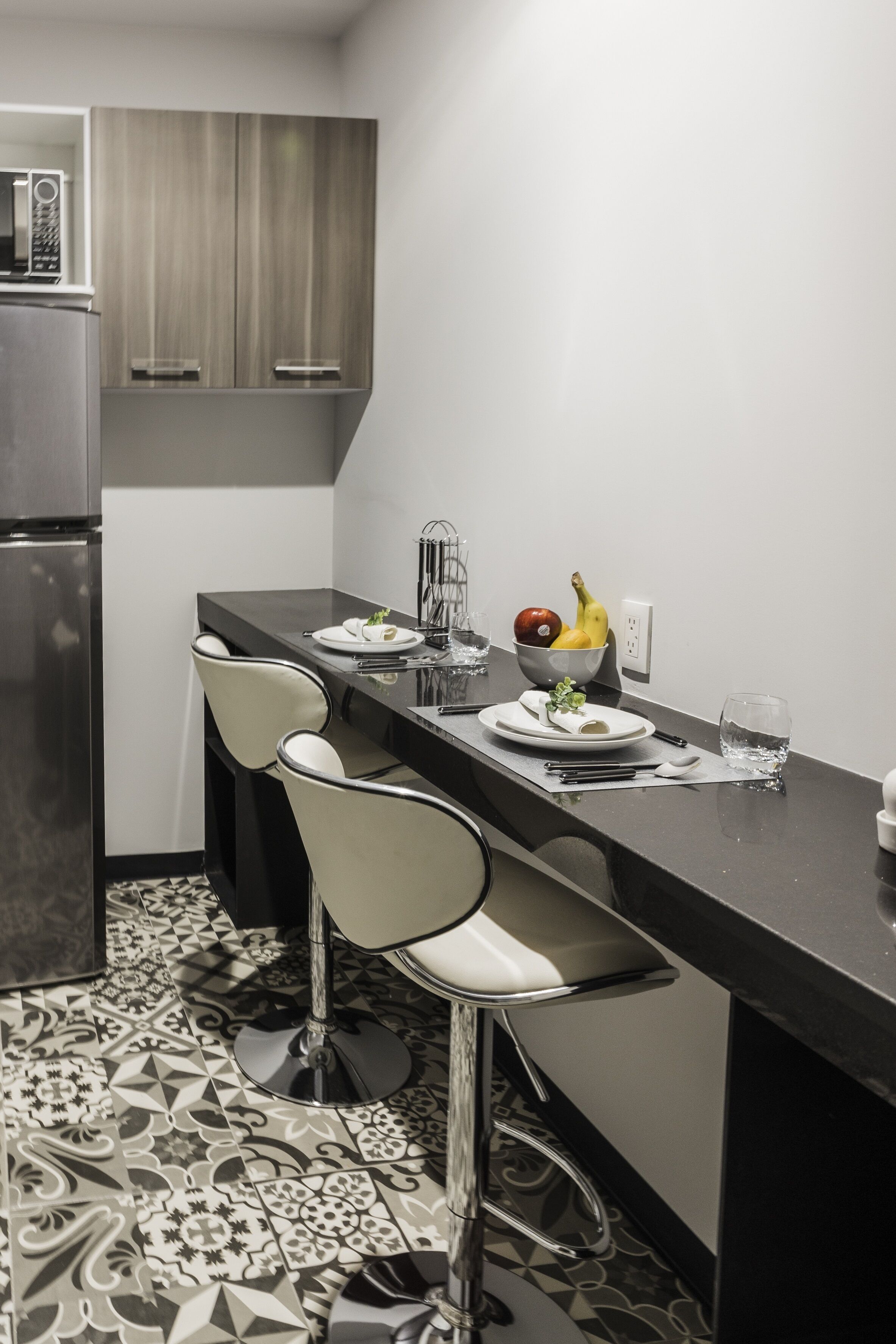 A modern kitchen with two bar stools, a dining table set with plates, a fruit bowl, and a microwave.
