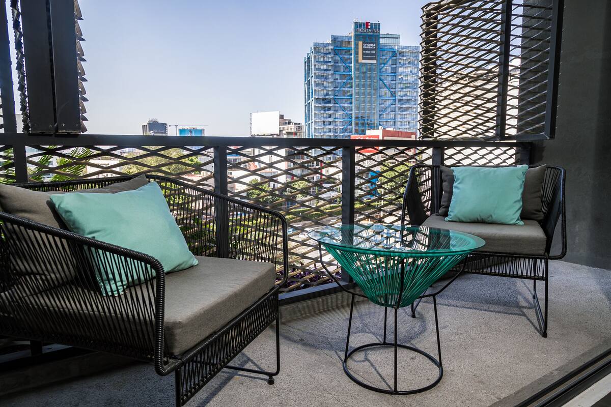 Un balcón con mobiliario de mimbre y una mesa de vidrio, con vista a un paisaje urbano de edificios modernos.