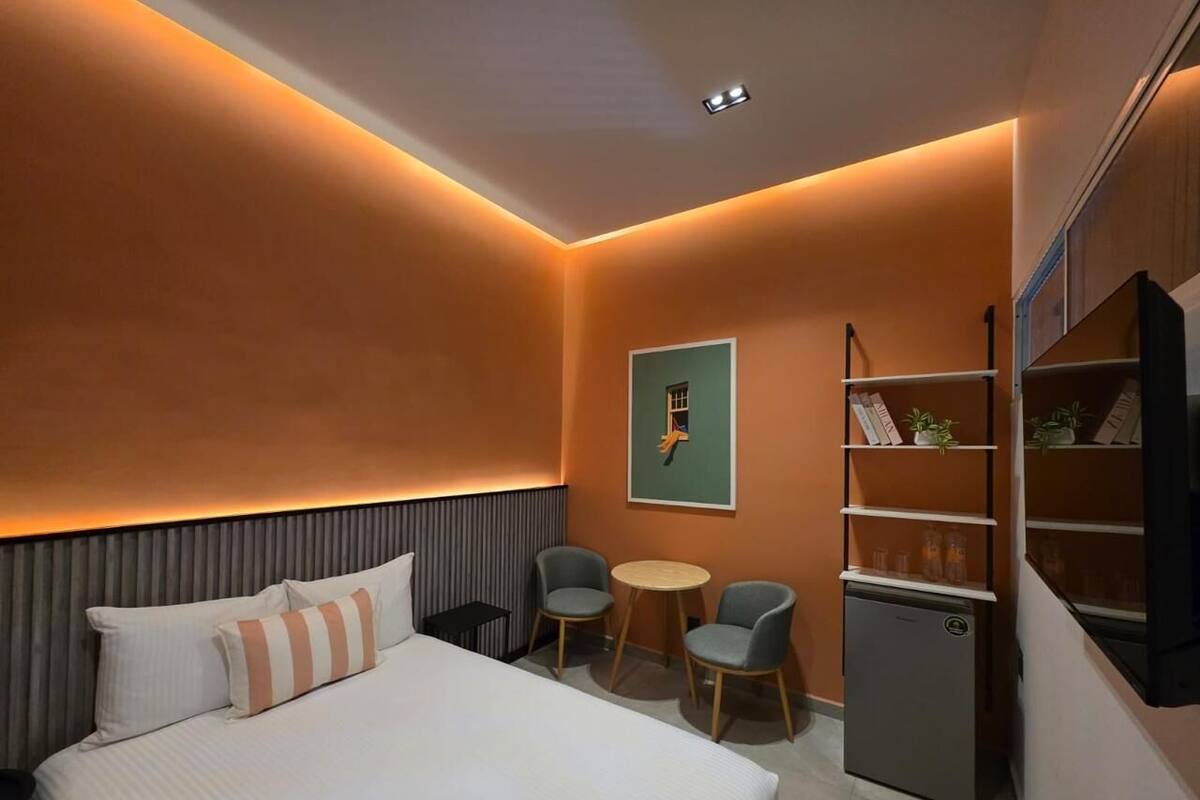 Habitación de hotel con una cama, dos sillas, una mesita, una estantería y un televisor.