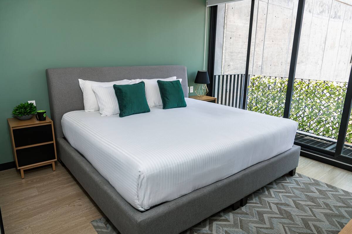 Un dormitorio con una cama grande, una mesita de noche, una planta en maceta y vistas al verde exterior.