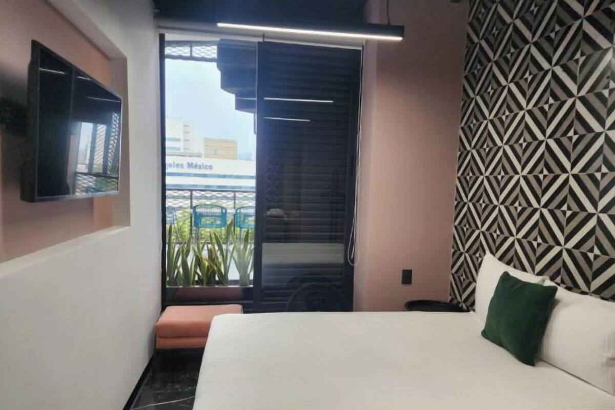 Habitación de hotel con cama, televisor montado en la pared, pared con diseño y ventana con vistas a edificios.