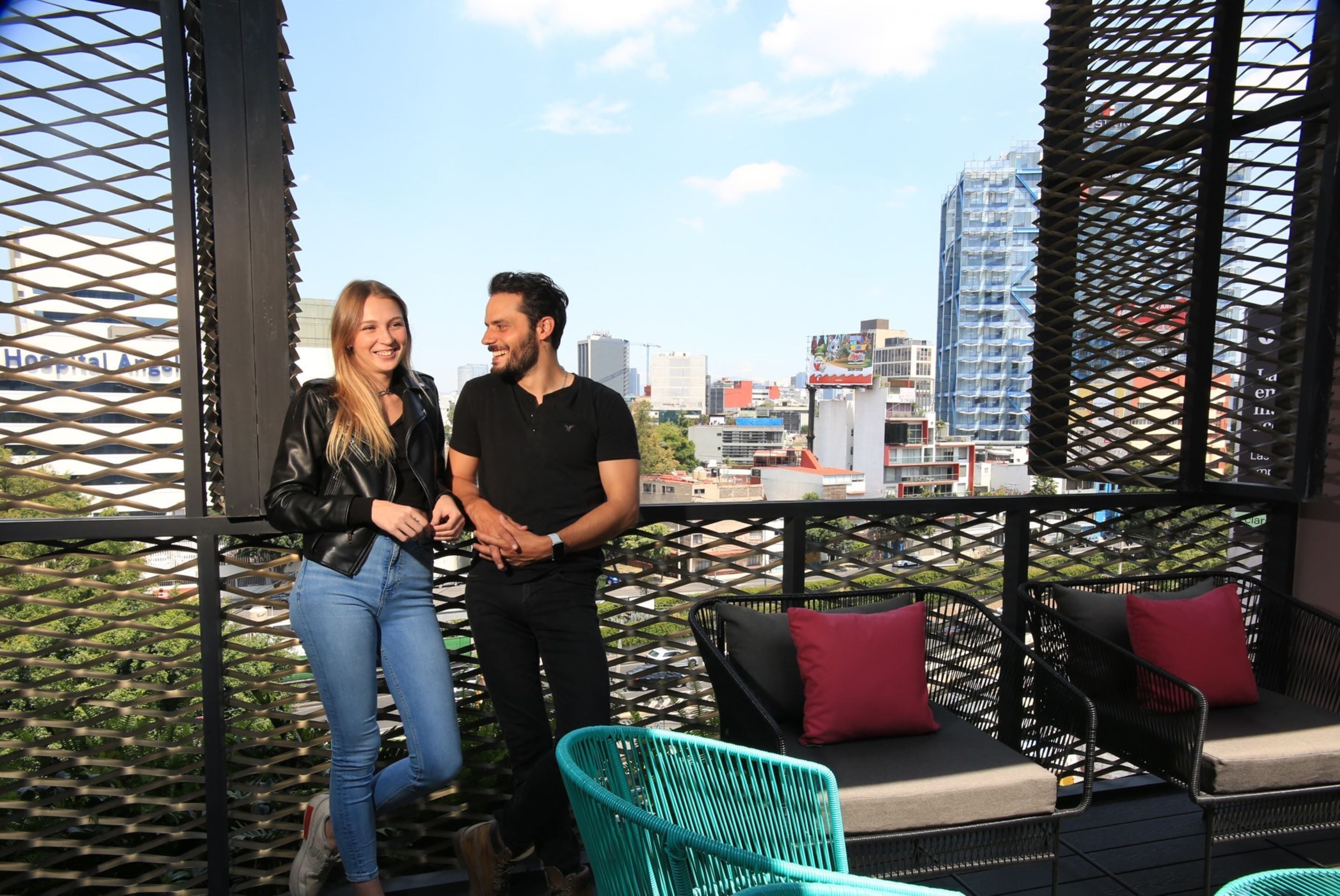 Een koppel staat op een balkon met uitzicht over de stad, met buitenmeubilair en een sierlijk rasterwerk.