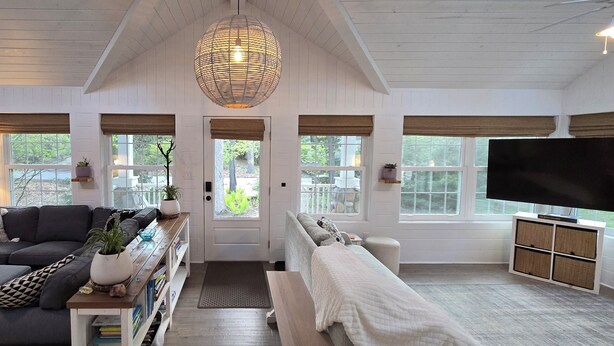 A spacious living room with a large sofa, a coffee table, and a television. There are large windows with blinds, a door, and a ceiling fan. - Relaxing Woodsy Cottage Retreat: Perfect for Families & Friends [Sleeps 8] (Michigan City)
