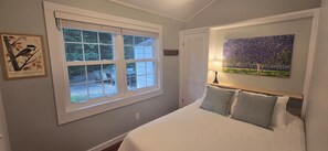 A bedroom with a bed, a window with blinds, a painting, and a chair outside. - Relaxing Woodsy Cottage Retreat: Perfect for Families & Friends [Sleeps 8] (Michigan City)