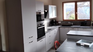 A modern kitchen with white cabinets, a built-in oven, a dining table with a red chair, and a large window providing natural light. - Charming gîte 300m from the picturesque village of Rougon (Rougon)