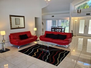Uma sala de estar moderna com dois sofás de couro vermelho, uma mesa de centro de vidro, um tapete com padrão preto e vermelho, uma obra de arte emoldurada na parede e uma área de jantar visível pela janela.