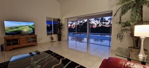 Sala de estar com televisão, sofá, mesa de centro e vista para uma piscina através de amplas portas de vidro.