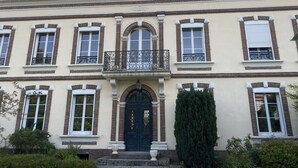 Um edifício de dois andares com fachada simétrica, destacando uma entrada central com porta preta, ladeada por janelas com persianas brancas.