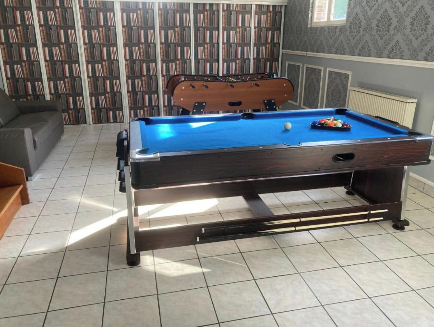 A pool table in a room with tiled flooring, a bookshelf in the background, and a radiator on the wall.