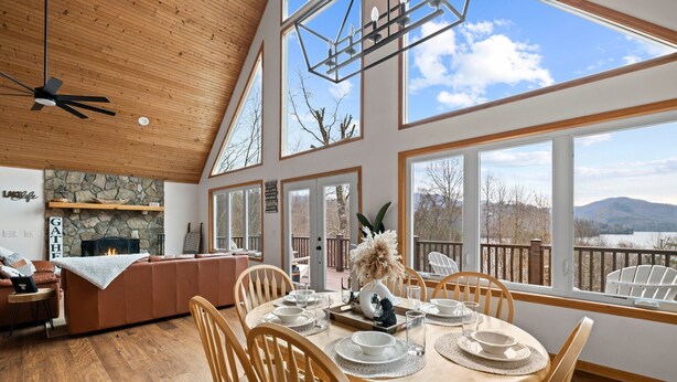 Un comedor amplio con una mesa de madera dispuesta para cuatro personas, rodeada de sillas de madera. Se observa una chimenea de piedra al fondo, y grandes ventanas ofrecen vistas al exterior.